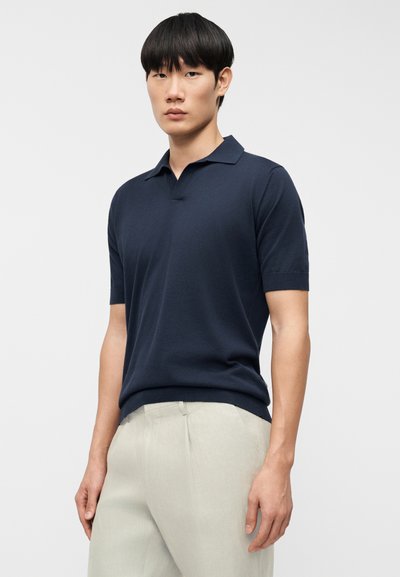 Polo de manga corta azul marino en material de punto suave con cuello clásico. Combinado con pantalones a medida en beige claro.