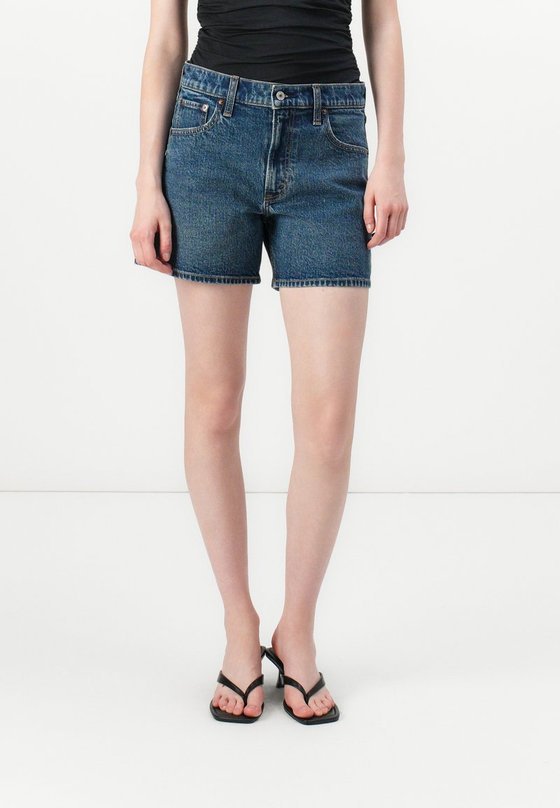 Donkerblauwe denimshorts met een klassieke snit, een tailleband, twee vinkzakken en een ruwe zoom. Vergezeld door zwarte slippers.