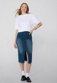 Mujer con una camiseta blanca de manga corta, falda midi de mezclilla azul con abertura frontal, calcetines blancos, mocasines negros y pendientes y pulsera de aro dorados.
