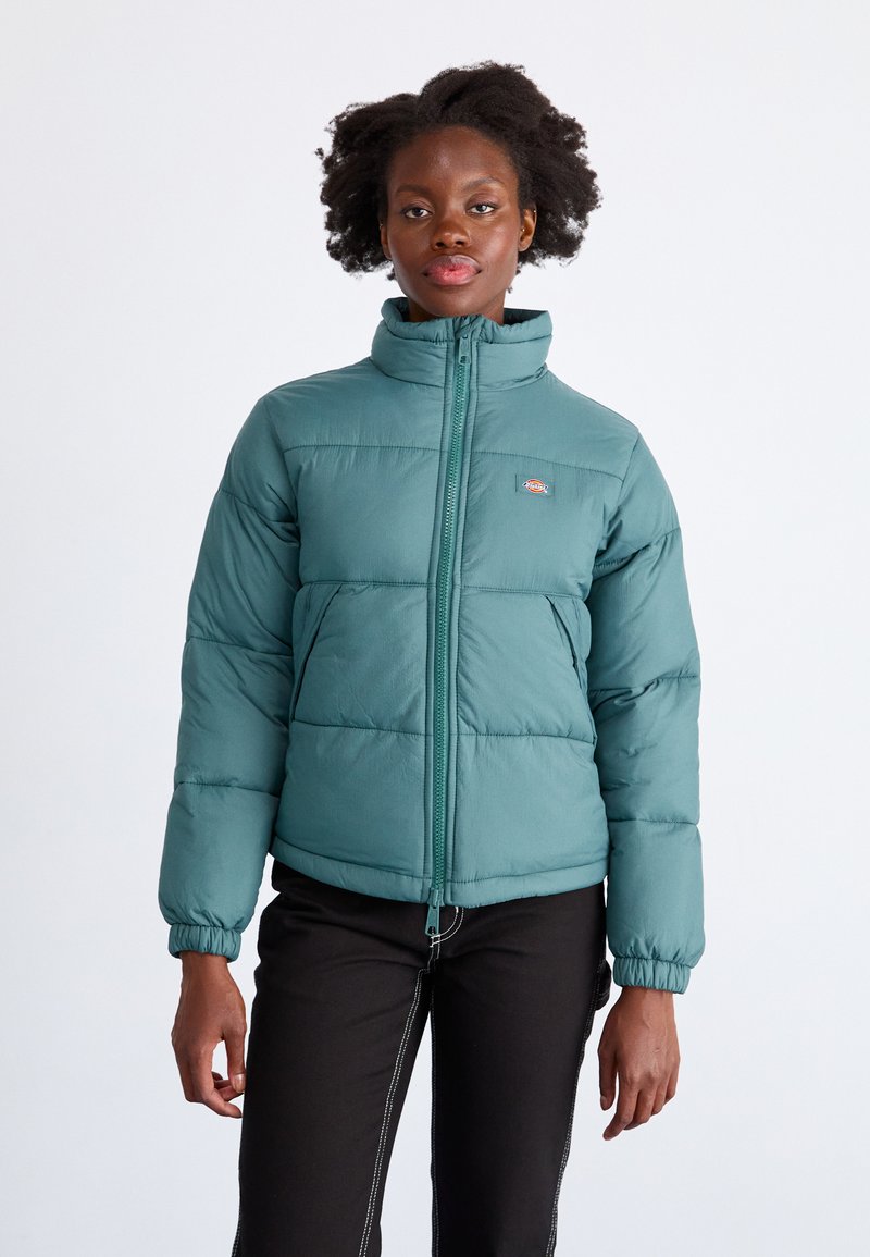 Tirkizna napihljena jakna visoke ovratnice, sprednjim zadrgo in elastičnimi manšetami. Ima kvadrirane panele in logotip na prsih.