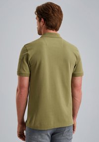 Olivinvihreä polo-paita, joka on valmistettu puuvillasta ja jossa on perinteinen kaulus ja lyhyet hihat. Vasemmassa hihassa on rintatasku.