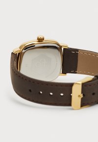 Reloj de color dorado con caja rectangular, correa de cuero marrón con costuras y hardware visible. La parte posterior está grabada con un logotipo.