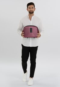 Purple hard-shell backpack with ridged design, black accents, and a front panel, held by a person in a white shirt and black pants.