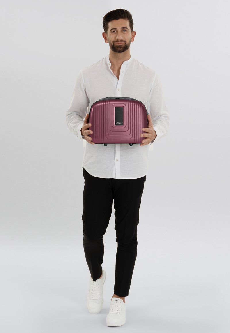 Purple hard-shell backpack with ridged design, black accents, and a front panel, held by a person in a white shirt and black pants.