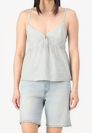 Woman wearing a light grey checkered sleeveless top with thin straps and light blue denim knee-length shorts, standing against a white background.