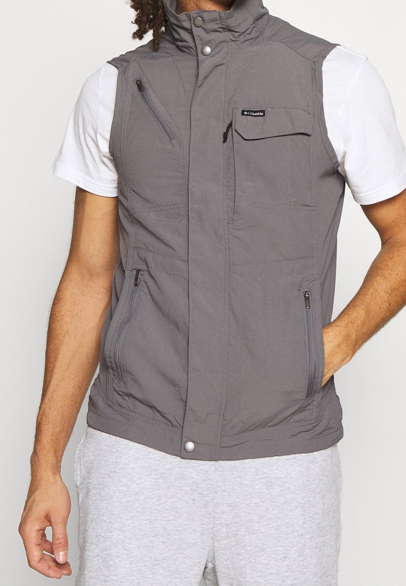 Gilet sans manches gris avec un col montant, multiple poches zippées et une texture lisse, arborant un logo dans la zone de la poitrine.