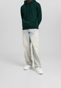 Grön hoodie med en framficka, kombinerad med ljusblå, något slitna jeans och vita sneakers. Enkelt design och avslappnad passform.