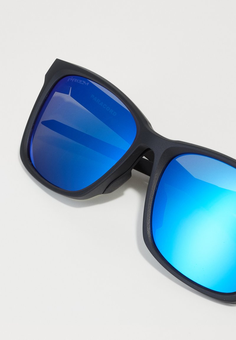 Occhiali da sole in plastica nera con lenti azzurro-tintate. Le lenti presentano una superficie riflettente e la montatura ha bordi angolari e una finitura opaca.