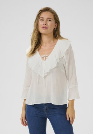 White blouse with a ruffled neckline and tie detail, featuring long sleeves with flared cuffs and a textured fabric. Worn with blue jeans.