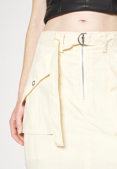 Taille et main d'une personne portant une jupe crème avec une fermeture éclair à l'avant, de grandes poches à rabat et une ceinture à boucle argentée, associée à un haut cropped noir.