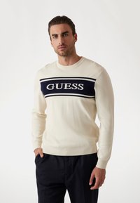 Pulover de culoare crem cu o bandă bleumarin pe care este inscripționat cuvântul "GUESS" în alb. Designul are un guler rotund și manșete și tiv cu nervuri.
