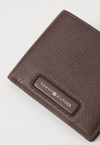 Portefeuille en cuir texturé marron avec des bordures cousues et un patch rectangulaire affichant le logo Tommy Hilfiger en blanc.