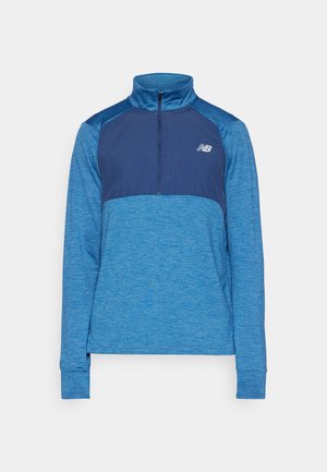 Sportovní tričko s dlouhým rukávem v různých odstínech modré. Obsahuje límec na zip do poloviny, texturovanou látku a logo na hrudi.