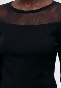 Gros plan d'une personne portant un top en maille noire avec un panneau en mesh transparent sur la partie supérieure de la poitrine et les épaules.