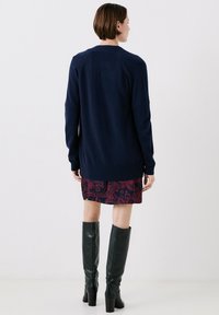 Maglione a maglia blu navy con maniche lunghe, abbinato a una gonna bordeaux con motivo. Il modello indossa stivali alti in pelle verde con tacco.