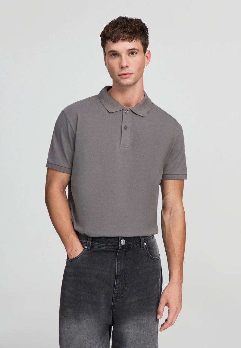 Młody mężczyzna w szarej koszulce polo z krótkim rękawem i ciemnych jeansach, stoi z jedną ręką w kieszeni na jednolitym tle.