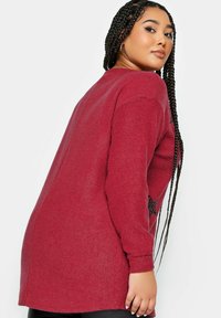 Röd räfflad stickad tröja med långa ärmar och avslappnad passform. Har svarta stjärndetaljer på sidorna. Slät textur, höftlång design.