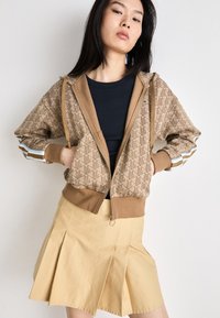 Bruine gemêleerde zip-up hoodie met een effen bruine zoom en elastische manchetten; gecombineerd met een beige geplooide rok.