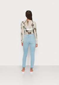 Top de mangas compridas, transparente, com um padrão marmoreado em cores suaves, combinado com jeans slim-fit azul claro. A modelo está de costas, usando chinelos.