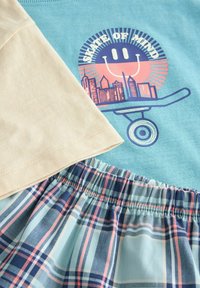 Ljusblå t-shirt med ett grafiskt tryck av ett leende ansikte och stadssilhuett på en skateboard. Äggskalsfärgade ärmar. Blå rutiga shorts med elastisk midja.
