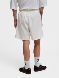 Άτομο που φοράει άσπρες σορτς με ελαστική μέση, άσπρες κάλτσες πλήρους μήκους με μαύρα ζιγκ ζαγκ και μαύρα αθλητικά Hummel, στέκεται μπροστά σε ένα μονόχρωμο φόντο.