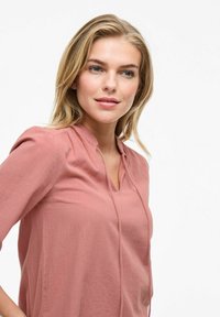 Dammig rosa blus med v-ringning och liten utskärning, med långa ärmar och ett mjukt, texturerat tyg.