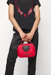 Červená prošívaná kabelka s černým srdíčkem a logem "LOVE MOSCHINO". Malý, obdélníkový tvar s horním uchem a zipovým uzavřením.