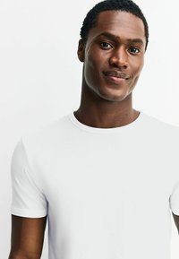 Balta īsroku T-krekls no gludas auduma ar apaļu kakla izgriezumu. Raksturīga pieguloša forma, minimālas šuves un nav redzamu rakstu vai grafikas.