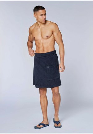 Muskulöser Mann, barfuß stehend, trägt einen marineblauen Handtuchrock mit Vordertasche und marineblaue Flip-Flops, schaut nach links vor einem einfarbigen Hintergrund.