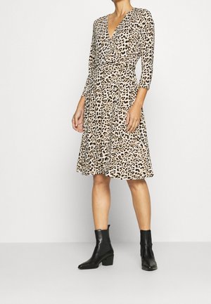 Frau trägt knieumspielendes, langärmliges Wickelkleid mit Leopardenmuster und V-Ausschnitt sowie schwarze Stiefeletten, steht vor einfarbigem Hintergrund.