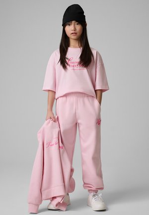 Młoda osoba nosi różowy, oversize t-shirt i spodnie dresowe z czerwonym napisem, trzymająca pasującą różową bluzę i czarną czapkę.