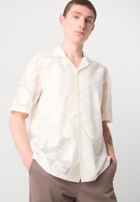 Chemisă cu mânecă scurtă, cu nasturi, din material subțire de culoare albă-crem, cu un model floral, croi lejer și guler. Asortată cu pantaloni maro deschis.