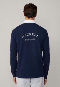 Mornarska polo majica dugih rukava s bijelim ovratnikom. Na leđima se nalaze natpisi "HACKETT" i "LONDON" u bijelom tekstu. Glatka tekstura tkanine.