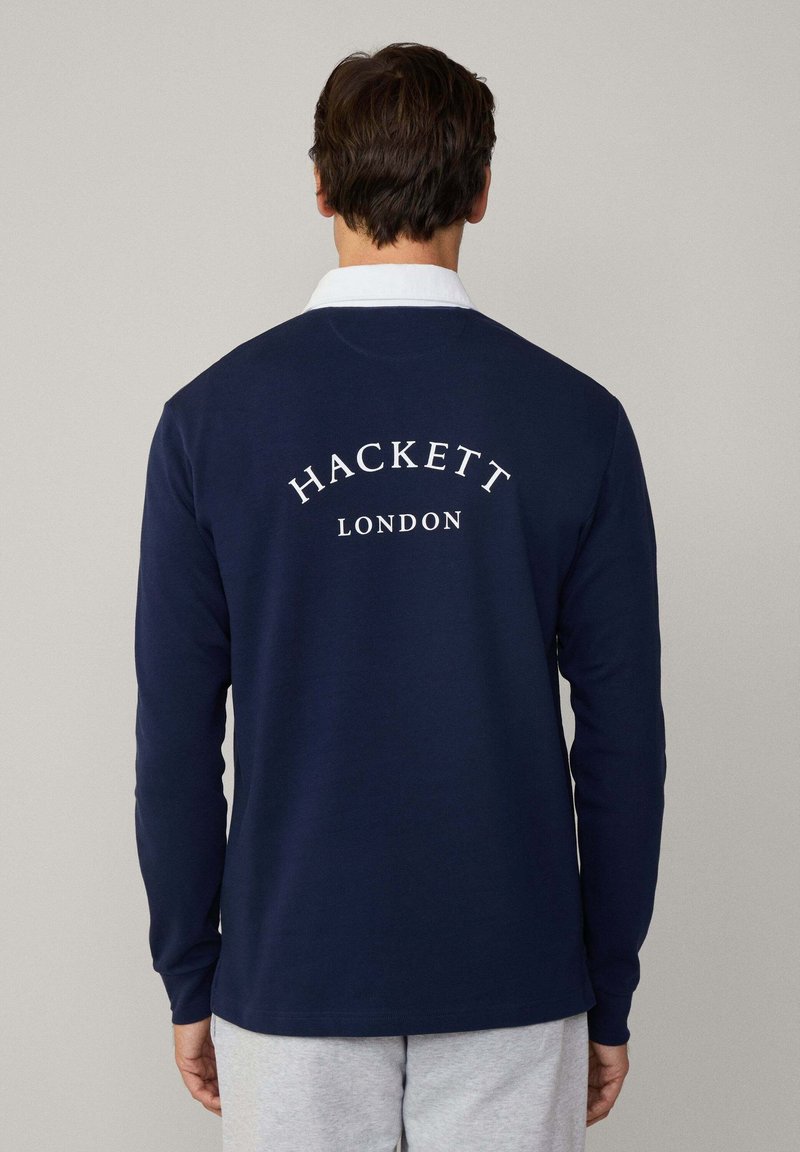 Mornarska polo majica dugih rukava s bijelim ovratnikom. Na leđima se nalaze natpisi "HACKETT" i "LONDON" u bijelom tekstu. Glatka tekstura tkanine.