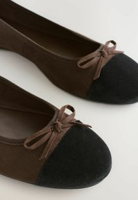 Braune Wildleder-Balletschuhe mit einer schwarzen Stoffzehenkappe; sie verfügen über eine Schleifenakzentuierung und einen elastischen Einstieg für einen sicheren Sitz.