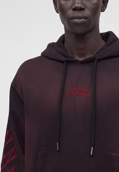 Homme portant un sweat à capuche sombre avec une broderie rouge "Off-White c/o AC Milan" sur la poitrine et un motif géométrique rouge sur la manche gauche.