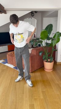 T-shirt branca com a estampa "Aroma do Verão", jeans cinzentos e ténis prateados. Ambiente interior com uma planta em vaso e um sofá castanho.