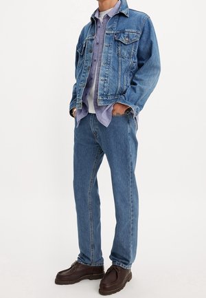 Man draagt een blauwe spijkerjack en spijkerbroek, lichtpaarse blouse met knopen, wit T-shirt en donkerbruine leren schoenen, staat met handen in de zakken.