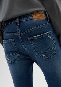 Tmavě modré džíny z denimu s pohledem zezadu, opotřebovanou texturou, jemným poškozením a dvěma zadními kapsami. Na pase je viditelná značka.