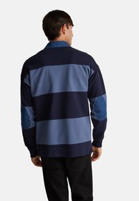 Tmavě modrá a světle modrá pruhovaná košile s dlouhým rukávem a denimovými nášivkami na loktech. Hladká textura, límečkový design a uvolněný střih.