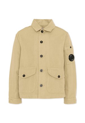 Giacca beige con bottoni, collo, due tasche frontali con patta, maniche con risvolto e una toppa circolare nera sulla tasca superiore del braccio sinistro.