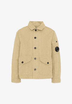 Giacca beige con bottoni, collo, due tasche frontali con patta, maniche con risvolto e una toppa circolare nera sulla tasca superiore del braccio sinistro.
