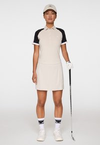 Beige golfklänning med kontrasterande svarta ärmar, korta muddar och en figursydd midja. Tillbehör inkluderar en keps, handskar och mönstrade strumpor.