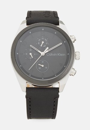 Montre avec bracelet en cuir noir, boîtier en acier inoxydable argenté, lunette texturée, cadran noir et index et aiguilles blancs.