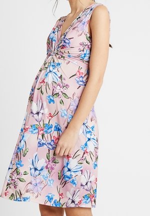 Zwangere vrouw in een mouwloze roze jurk met een blauw-wit bloemenpatroon, zijkantprofiel van schouders tot knieën.