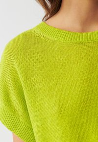 Detailní záběr na osobu v jasně limetkově zeleném pleteném svetru s krátkým rukávem a vroubkovaným kulatým výstřihem.