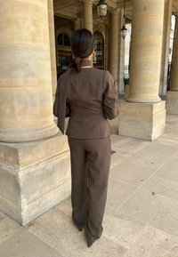 Hnedý strihový oblek so štruktúrovanou bundou a širokými nohavicami, vyrobený z hladkej tkaniny. Stojí pred kamennými stĺpmi na námestí.