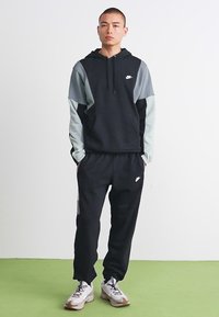 Man wearing black and grey Nike hoodie and black Nike sweatpants standing with hands in pockets on green floor.
