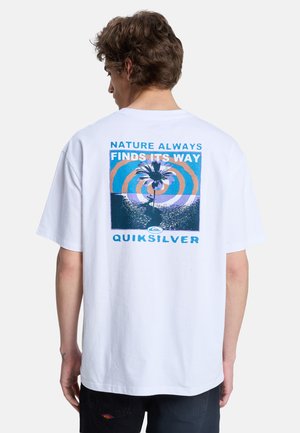 Jeune homme aux cheveux bruns courts portant un t-shirt blanc avec un graphisme d'arbre de palmier et le texte « Nature Always Finds Its Way » ainsi que « Quiksilver » au dos.