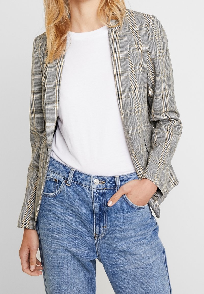 Personne portant un blazer à carreaux gris sur un t-shirt blanc et un jean bleu taille haute, une main dans la poche et l'autre détendue.